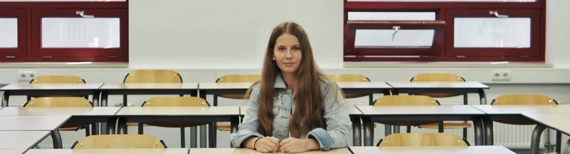 Portretfoto van Annemae de Vries, ze zit in een klaslokaal aan een tafel op een stoel. Haar lange donkerblonde haar zit los, ze draagt een lichtblauw spijkerjasje, witte jeans en witte sneakers