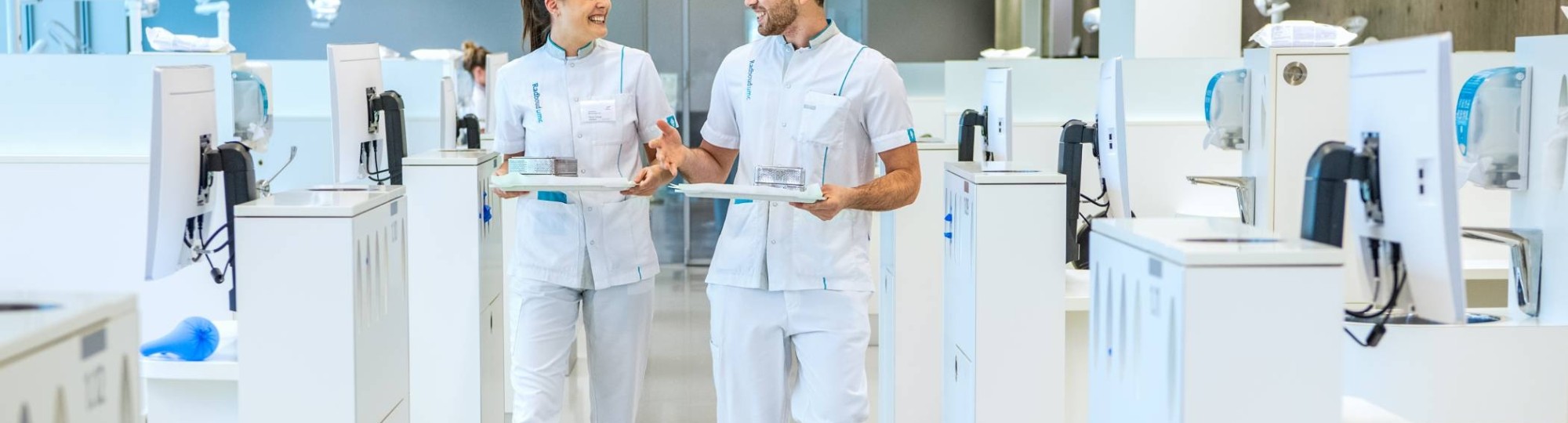 2 studenten kijken elkaar aan en lopen door een ruimte met veel behandelstoelen.