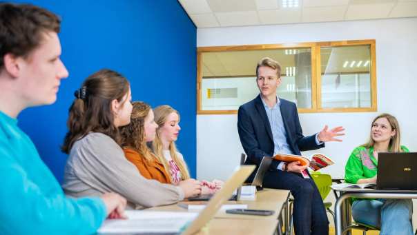 Docent in donkerblauw pak zit op de tafel met een boek in zijn hand, terwijl hij zijn studenten uitleg geeft
